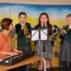 Three students playing trumpet and clarinet while their teacher plays piano