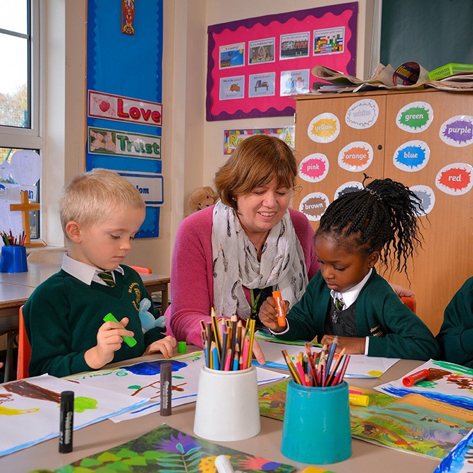 Two pupils colouring in at their desk with a teacher looking over them