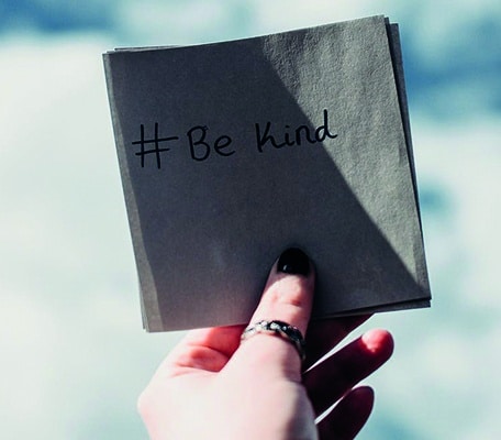 A woman's hand holding a slip of paper with the words # Be Kind written in pen