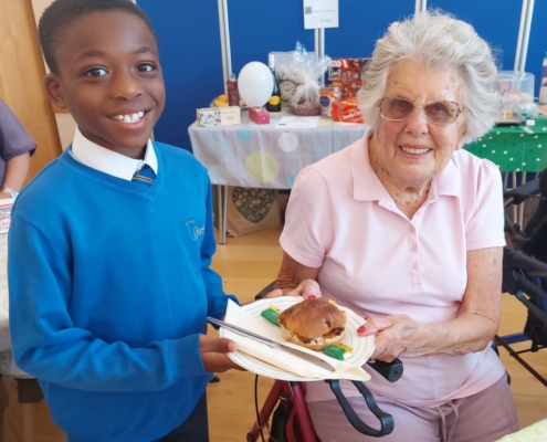 Pupils smiling and handing a plate of cake over to an elderly lady