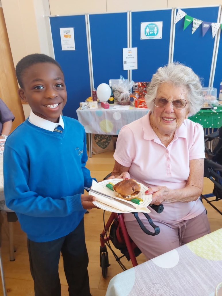 Pupils smiling and handing a plate of cake over to an elderly lady